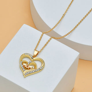 Gold-toned heart-shaped pendant necklace with "Mom" text and diamond accents, displayed on a white surface against a soft orange background.