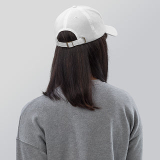 White baseball cap on long dark hair on woman's head and upper back in gray sweatshirt against plain background.