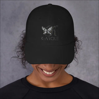 Black dad hat with K-AROLE logo in a minimalist style, worn by a smiling person against a plain gray background.