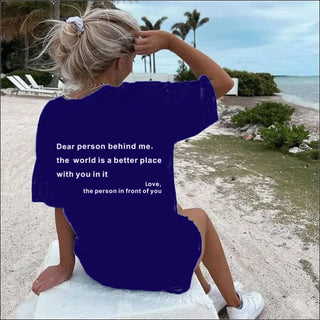 Woman on a beach with sandy shoreline and palm trees in the background wearing a navy blue T-shirt with a motivational message printed on it.